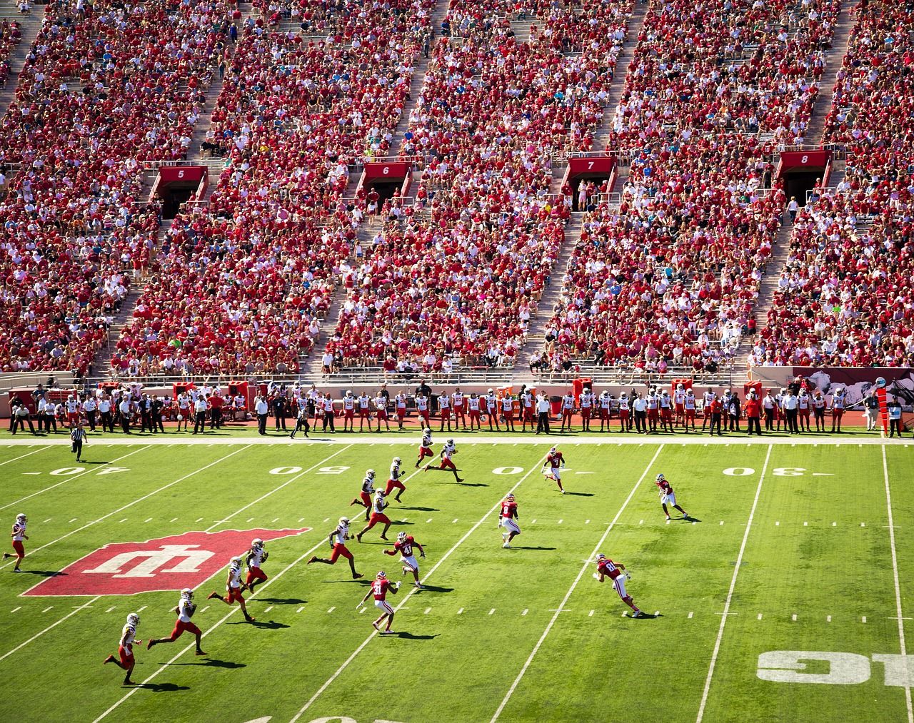 Vista aerea di una partita di calcio americano affollata all'Università dell'Indiana, con giocatori in azione sul campo e tifosi sugli spalti.