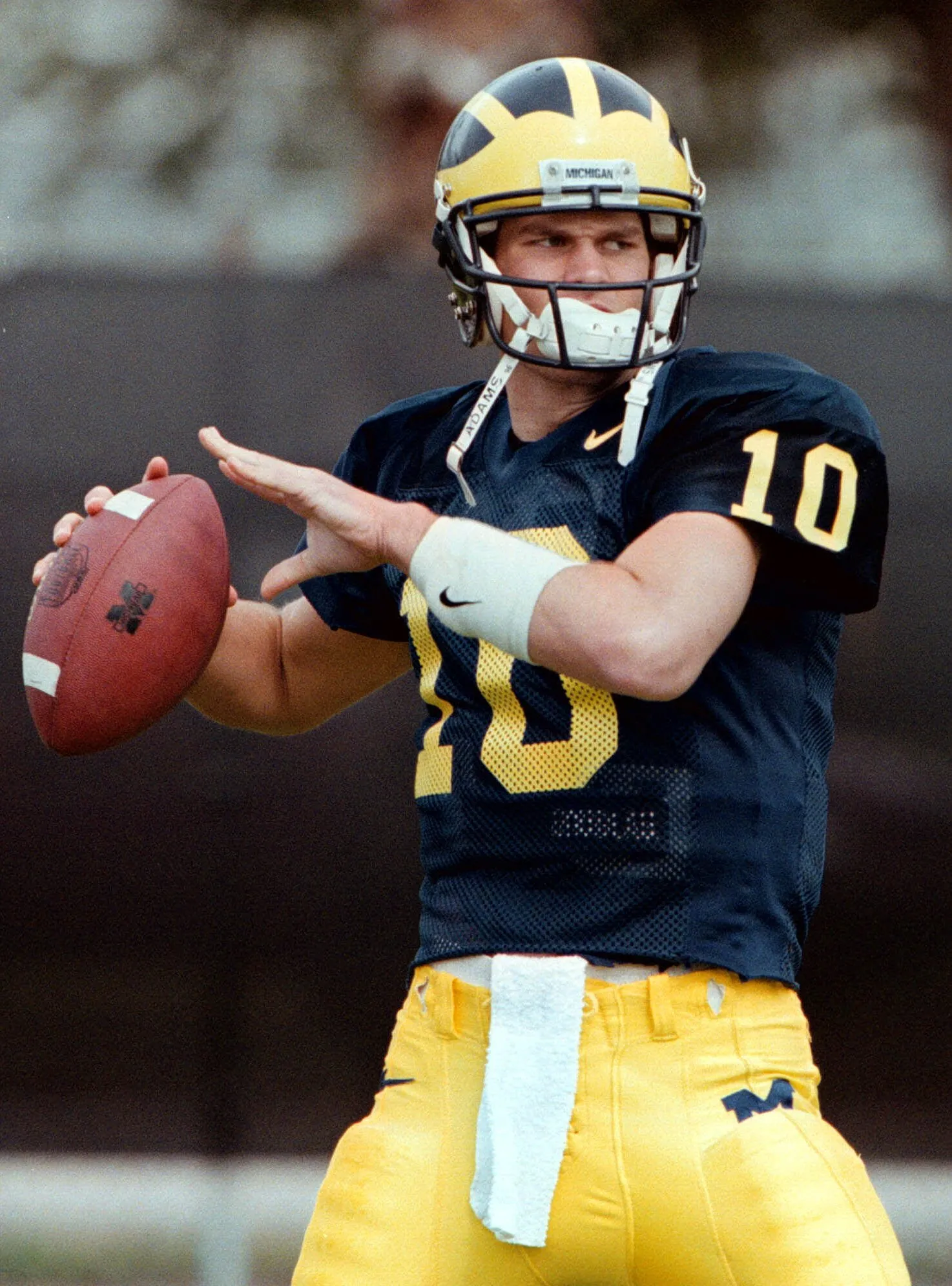 Giovane Tom Brady in azione come quarterback per l'Università del Michigan durante una partita di football universitario, vestito con l'uniforme blu e gialla della squadra.